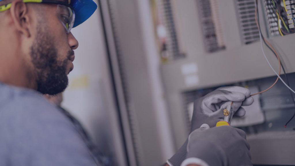 African-American electrician with a hardhat and protective eyewear stripping a wire while wearing protective gloves.